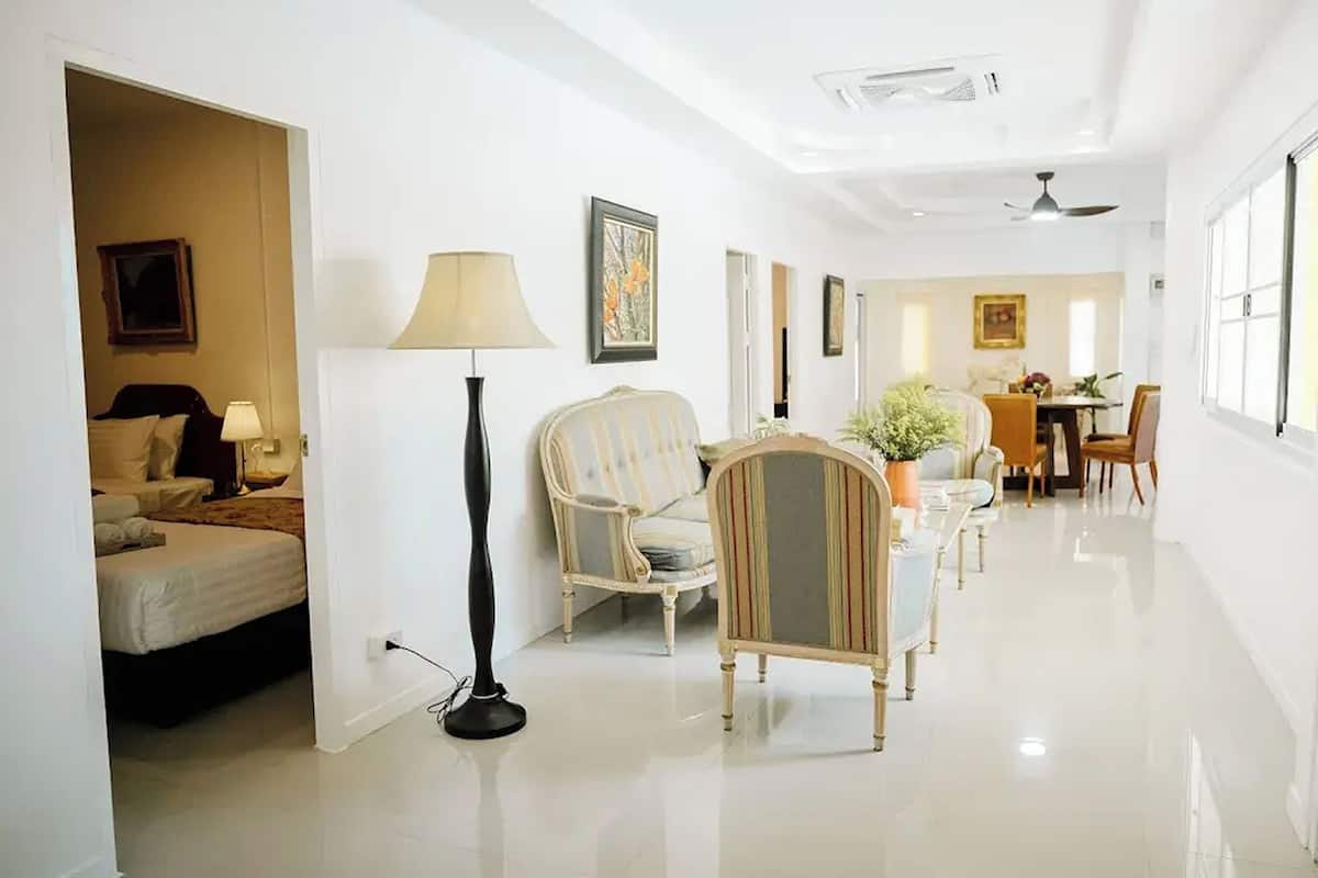 A hotel room with a bed, a standing lamp, a striped armchair, and a dining area with a table and chairs.