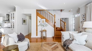 Ein geräumiges Wohnzimmer mit einer Holztreppe, weißen Wänden und einer Küche im Hintergrund.