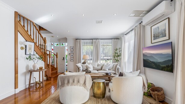 Ein geräumiges Wohnzimmer mit einer Holztreppe, einer Sofagarnitur, einem Couchtisch und einem großen Fenster mit Vorhängen.