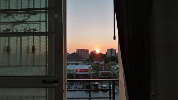 A view through a window with metal bars, a sunset, and buildings in the background. - Eco hostel Bangsaen (Chon Buri)
