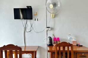 Ein Zimmer mit einem an der Wand befestigten Flachbildfernseher, einem Standventilator, einem Holztisch und einem Stuhl.