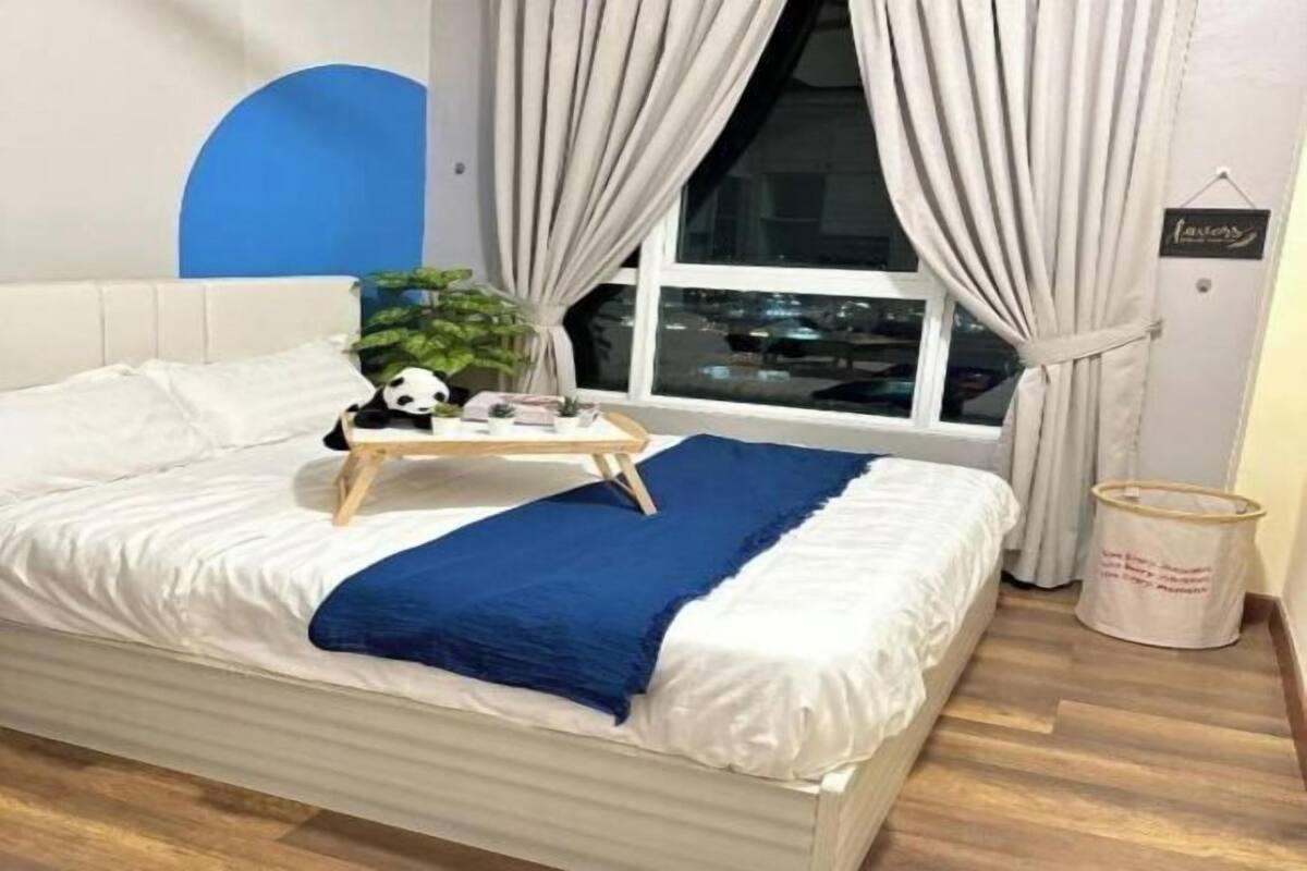 Cama bem arrumada com um cobertor azul, uma pequena bandeja de madeira com um brinquedo de panda e uma planta, e uma janela com cortinas.