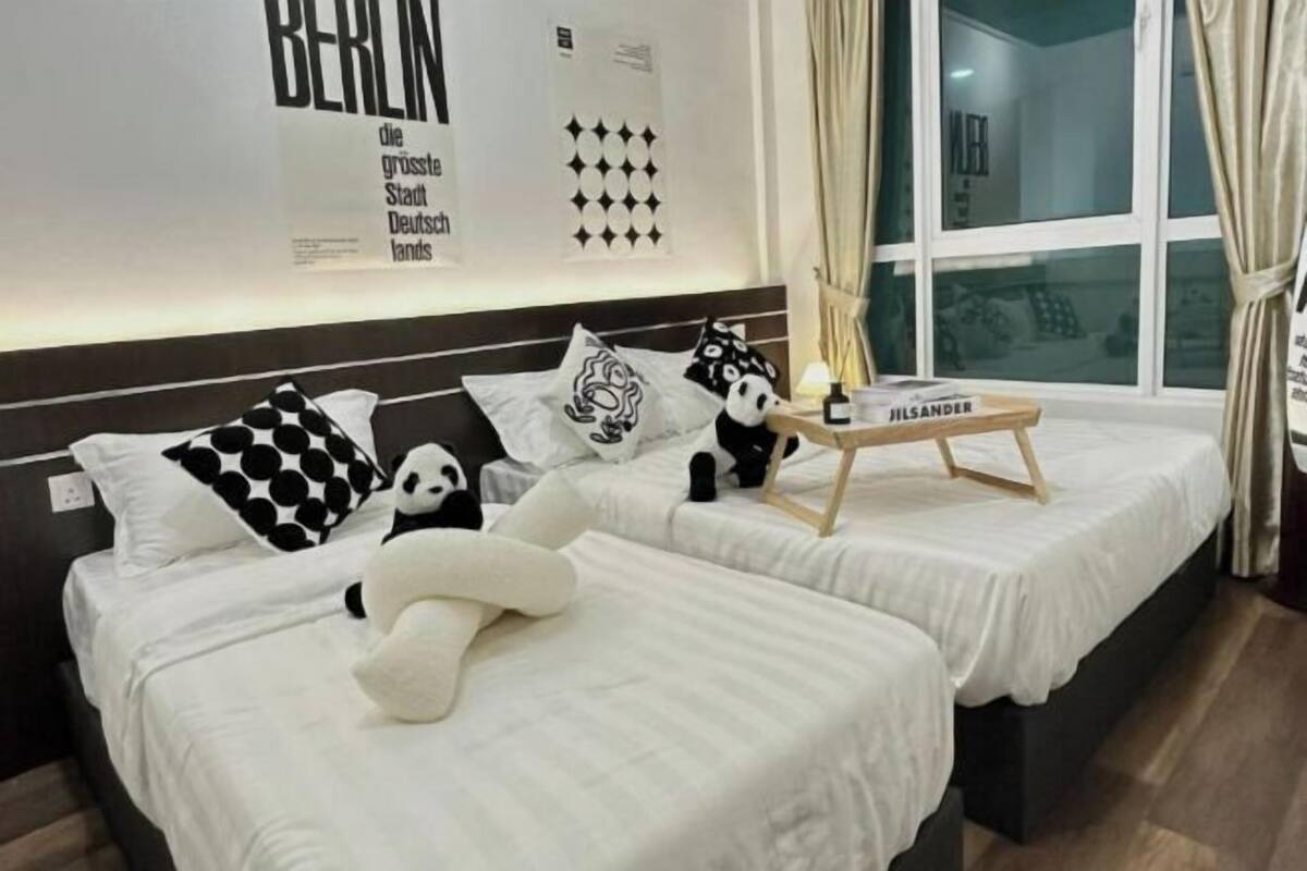 Quarto de hotel com duas camas, cada uma com colcha branca e almofadas estampadas em preto e branco. Há uma pequena mesa de madeira com uma luminária e uma garrafa com a etiqueta "HILSANDER". O quarto tem uma janela grande com cortinas, e a parede apresenta um pôster com o texto "BERLIN die größte Stadt Deutschlands".
