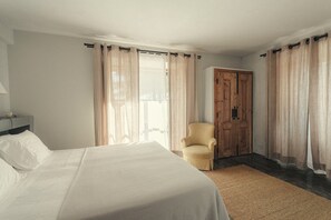A hotel room with a bed, a chair, a wooden door, and a window with curtains.