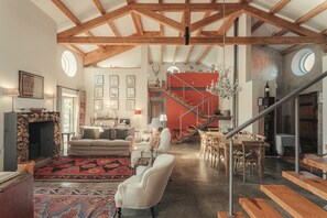 A spacious living area with a high ceiling, a fireplace, a dining table, and a staircase leading to an upper level.