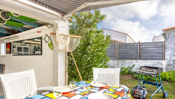 Une terrasse avec une table dressée pour les repas, des chaises d’extérieur, un chariot-barbecue et une grande plante.