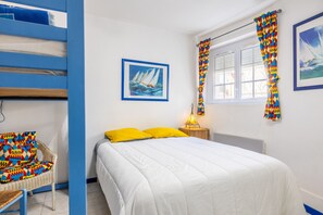 A bedroom with a bed, a chair, a painting, a window with curtains, and a nightstand with a lamp.