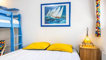 A room with a bunk bed, a painting of sailboats, a wooden nightstand with a lamp, and a colorful curtain.