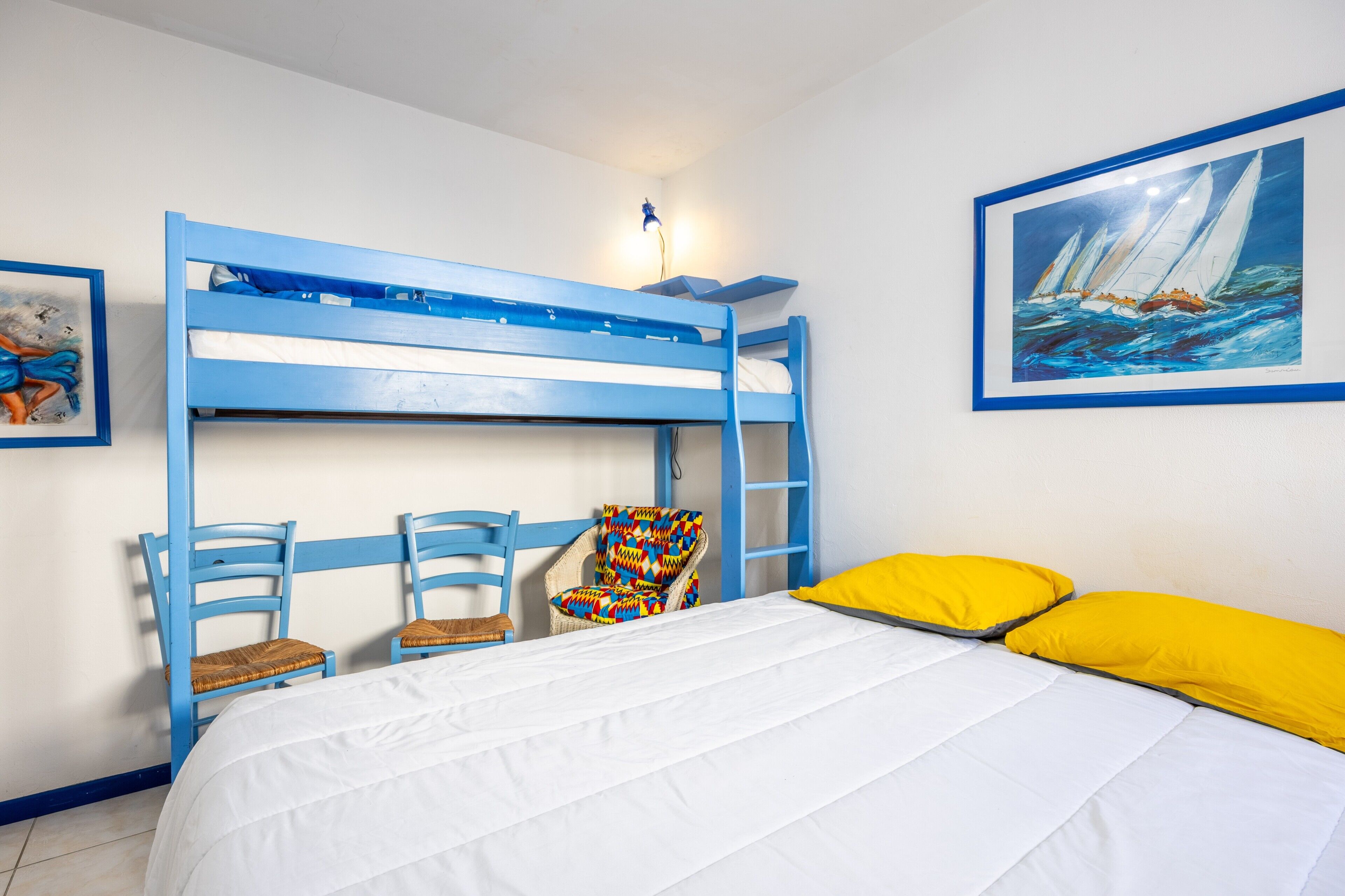 Une chambre avec un lit superposé, un matelas blanc, deux chaises bleues, une chaise colorée, un tableau encadré et un coussin jaune.