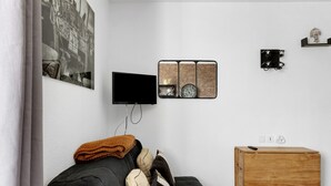 A modern living room with a black sofa, a wooden side table, a flat-screen TV, and a wall-mounted clock. - Apartment 'L'aventurier' – Steps from Futuroscope, Sleeps 4 (chasseneuil du Poitou)
