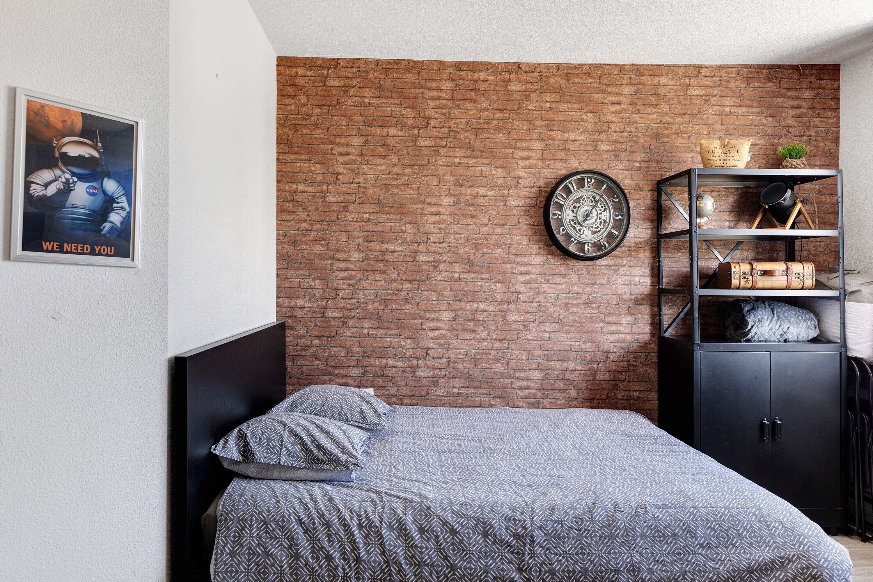 A bedroom with a brick wall, a bed with patterned bedding, a clock, a shelf with various items, and a poster on the wall.