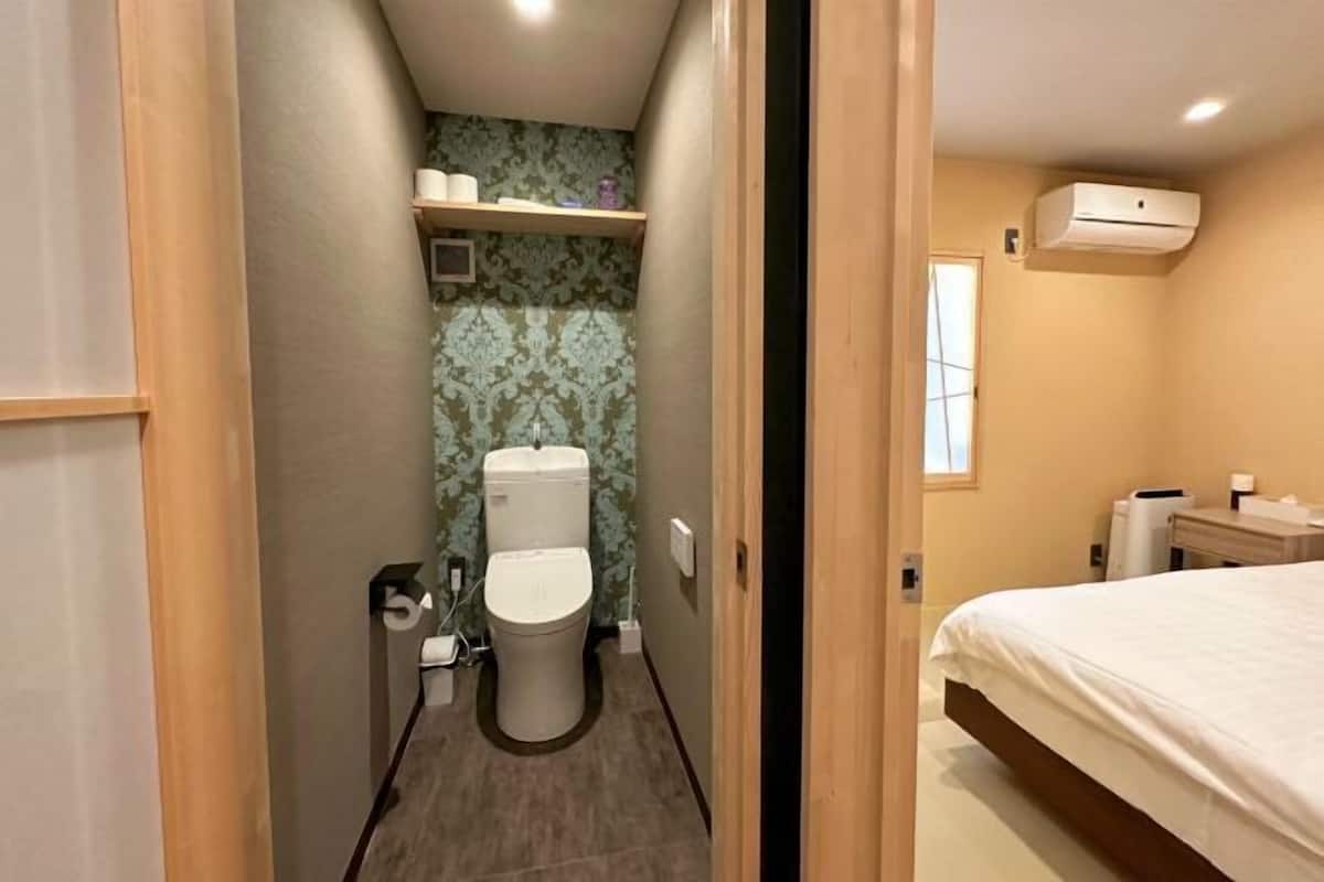 A narrow hallway leading to a bathroom with a toilet, a shelf with toiletries, and a bed with a white bedspread.