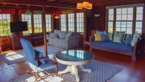 Una sala de estar acogedora con un sofá azul, un reposapiés a rayas y una mesa redonda. Amplias ventanas ofrecen vistas al exterior.