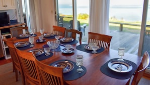 Una mesa de comedor con platos azules y blancos, vasos y cubiertos, con vista al mar a través de amplias puertas correderas.