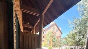 Un espacio de estar al aire libre cubierto con mobiliario de madera, pared de piedra y vistas a un edificio y árboles.
