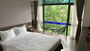 A hotel room with a large bed, a small table, a chair, and a balcony with a view of greenery. - 3T Hotel Hanoi (Hanoi)