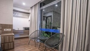 A hotel room with a wooden floor, two black chairs, a small table, a window with curtains, and a reception desk in the background. - 3T Hotel Hanoi (Hanoi)
