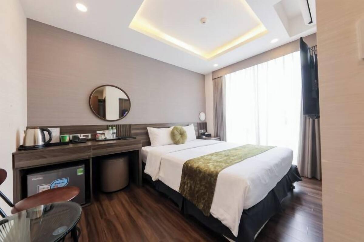 A hotel room with a bed, a desk, a chair, a small table, a mirror, a window with curtains, and a ceiling light fixture.