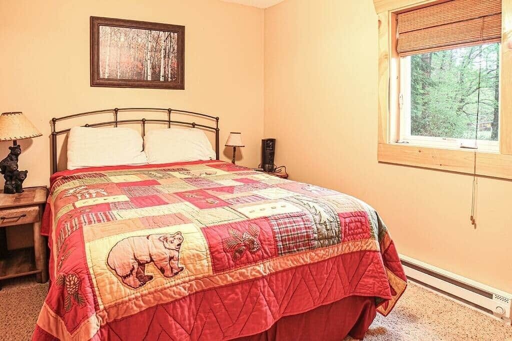 Un dormitorio con una cama grande, una ventana con vistas a árboles, una mesita de noche con lámpara y un cuadro en la pared.
