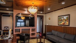 A modern living room with a large flat-screen TV, a grey sofa, a wooden ceiling, and a decorative chandelier. - The Ambassador's Terrace (Belize City)
