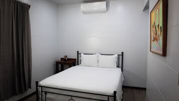 Un dormitorio con cama, aire acondicionado, mesita de noche, un cuadro y una ventana con cortinas.