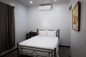 Un dormitorio con cama, aire acondicionado, mesita de noche, un cuadro y una ventana con cortinas.
