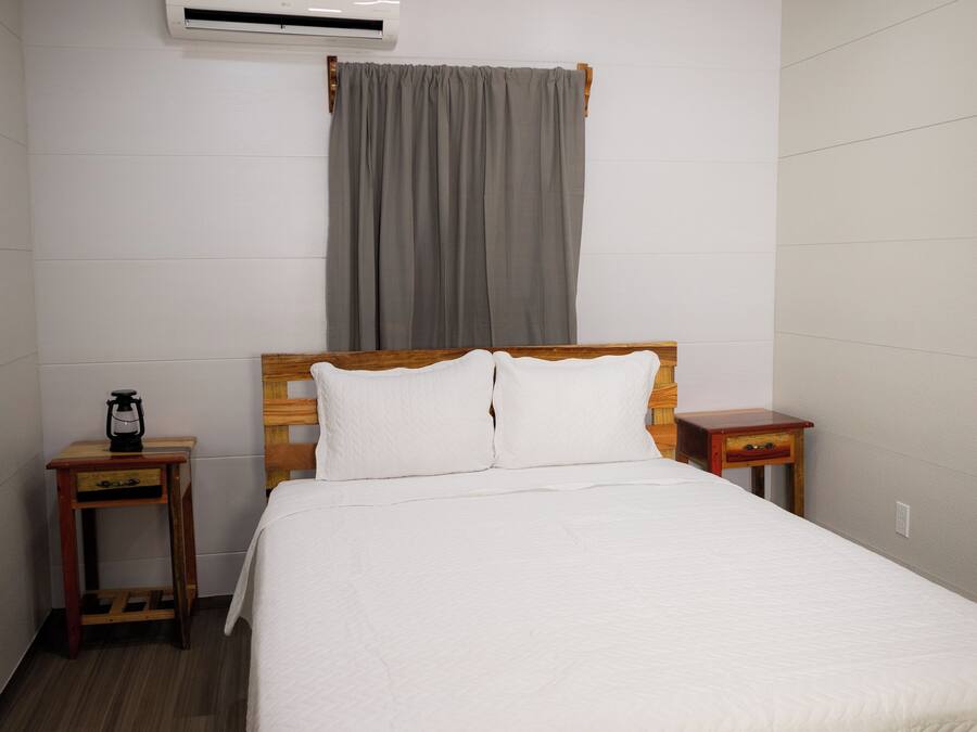 Cama bem arrumada com colcha branca, cabeceira de madeira e cortina cinza. Tem uma pequena mesa de cabeceira de madeira com uma luminária e um ar condicionado fixado na parede acima da cama.