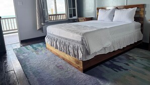 A bedroom with a wooden bed frame, white bedding, and a view of the ocean through an open door. - The Ambassador's Terrace (Belize City)