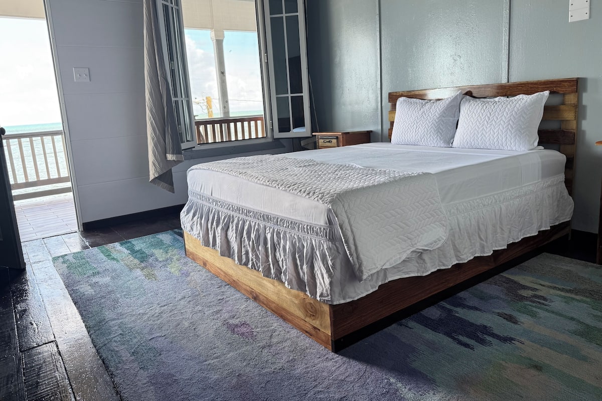 Ein Schlafzimmer mit einem hölzernen Bettgestell, weißer Bettwäsche und Blick auf das Meer durch eine offene Tür.