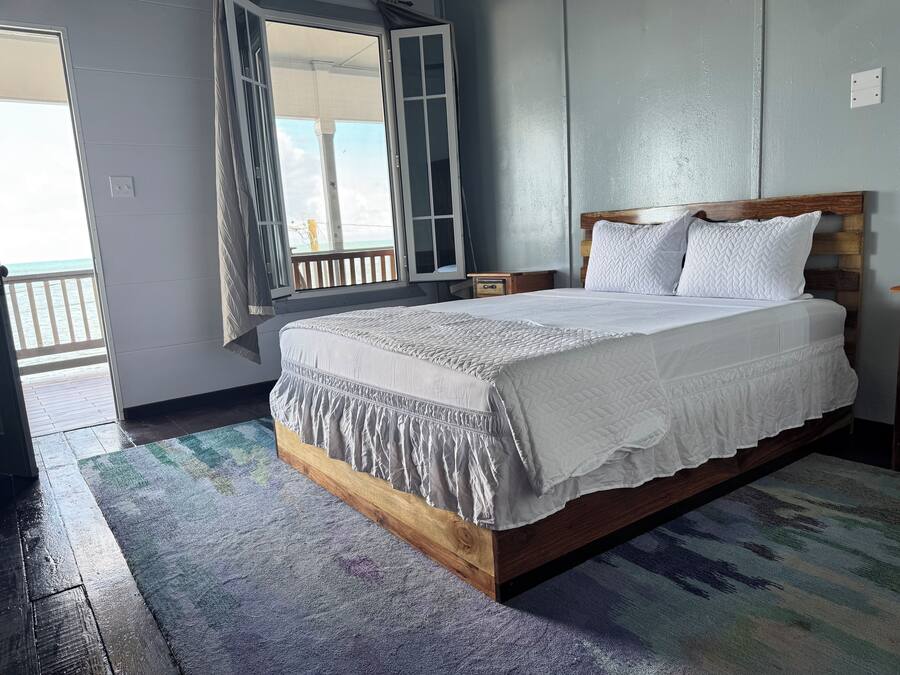 A bedroom with a wooden bed frame, white bedding, and a view of the ocean through an open door.