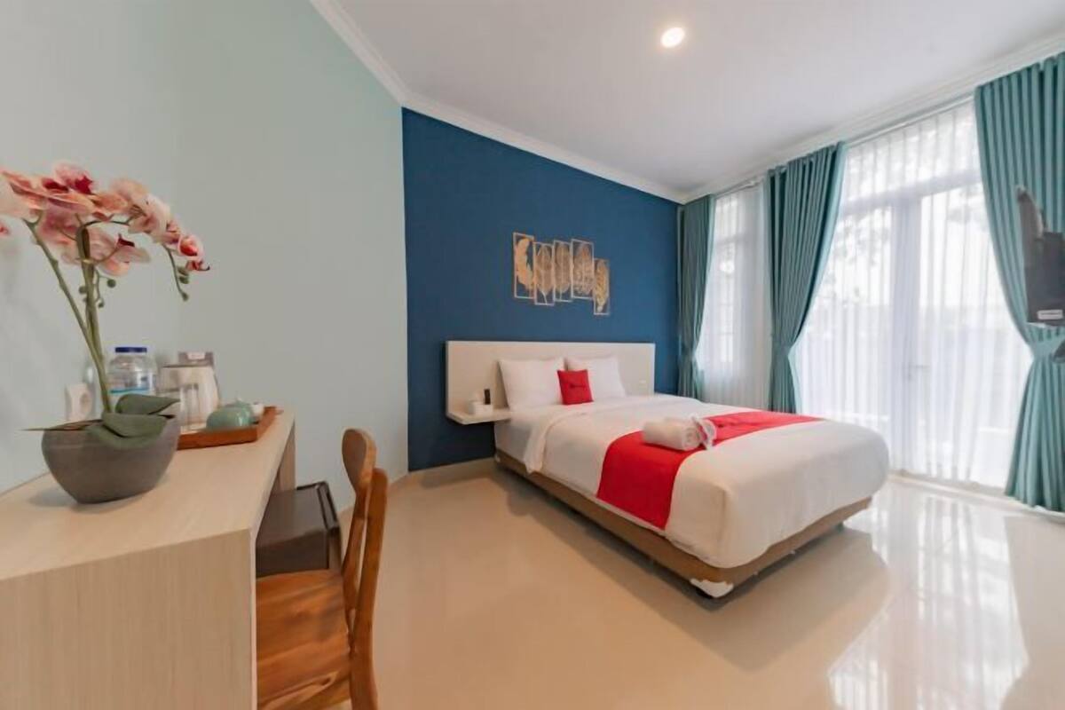 Habitación de hotel con cama, escritorio, silla, un florero con flores y una obra de arte en la pared.