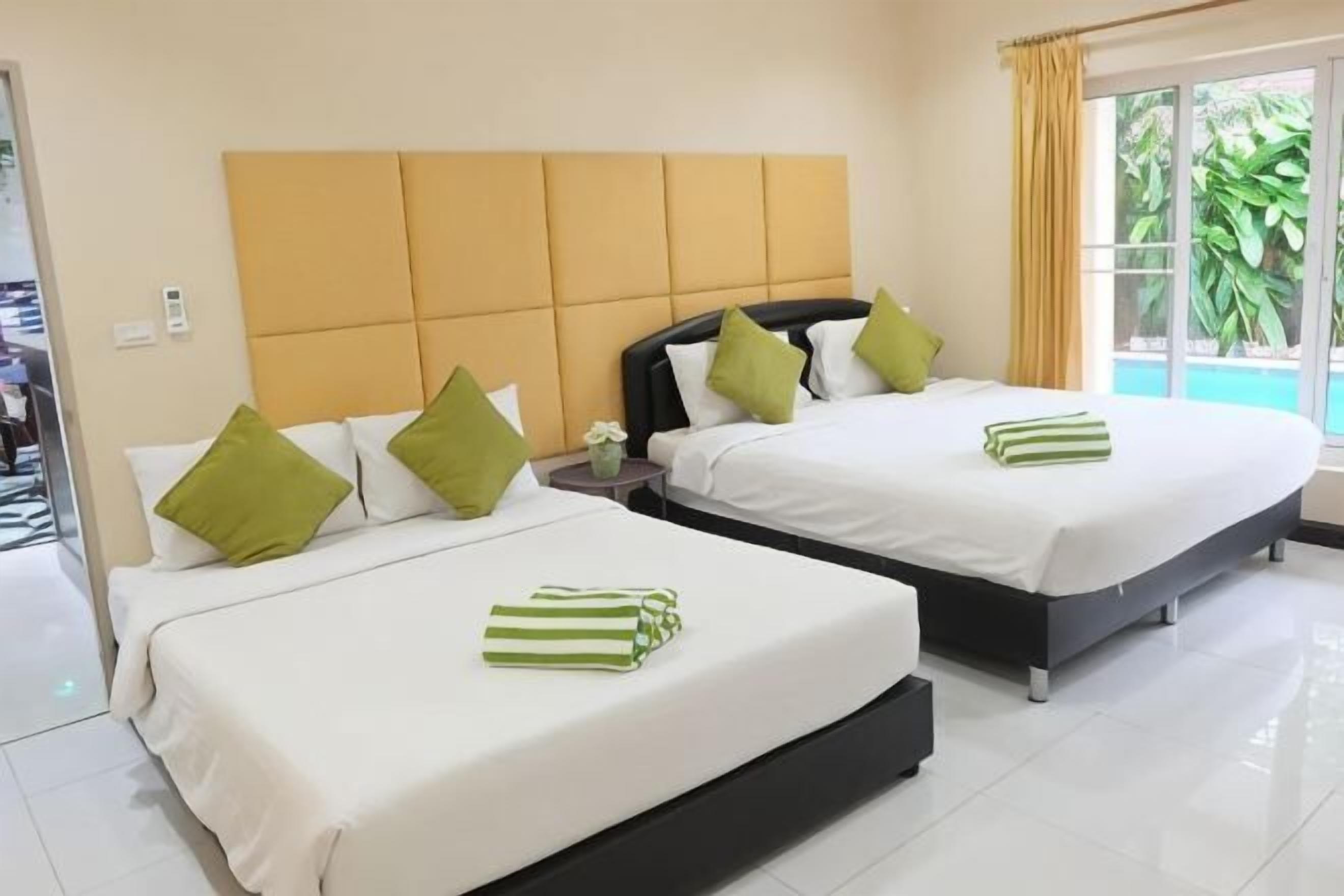 Duas camas com roupa de cama branca e almofadas verdes, uma cabeceira de madeira, uma mesa pequena com um vaso e uma janela com cortinas.