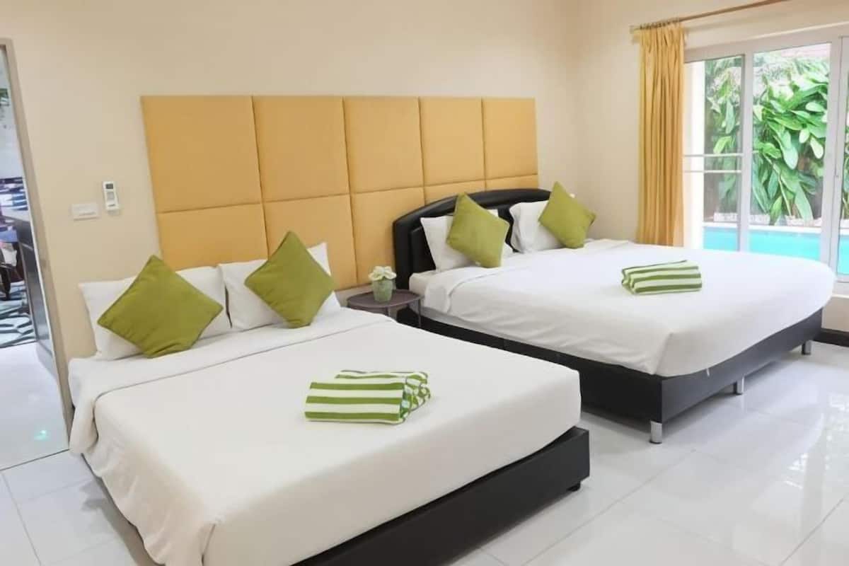 Two beds with white linens and green pillows, a wooden headboard, a small table with a vase, and a window with curtains.