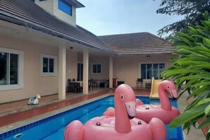 A swimming pool with inflatable flamingos, a patio area, and a house with a tiled roof. - The Pool House Pattaya No.2- 5 bedroom (Pattaya)