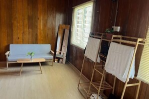A room with a wooden floor, a sofa, a small table with a plant, a drying rack with towels, and a window with blinds. - Manson Goat Cottage (A) - Nature escape hideaway (Ulu Telom)