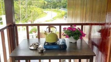 A wooden table on a balcony with a kettle, a portable stove, a pot, and a potted plant with pink flowers.