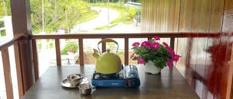 A wooden table on a balcony with a kettle, a portable stove, a pot, and a potted plant with pink flowers.
