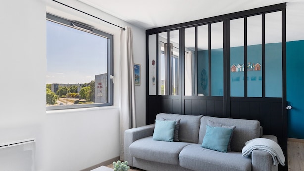 A modern living room with a grey sofa, a small table with a plant, and a view of the city through a large window.