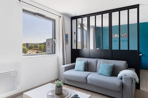 A modern living room with a grey sofa, a small table with a plant, and a view of the city through a large window. - Cabano'scope – Futuroscope 300 m, Cozy and Quiet for 4 Guests (chasseneuil du Poitou)