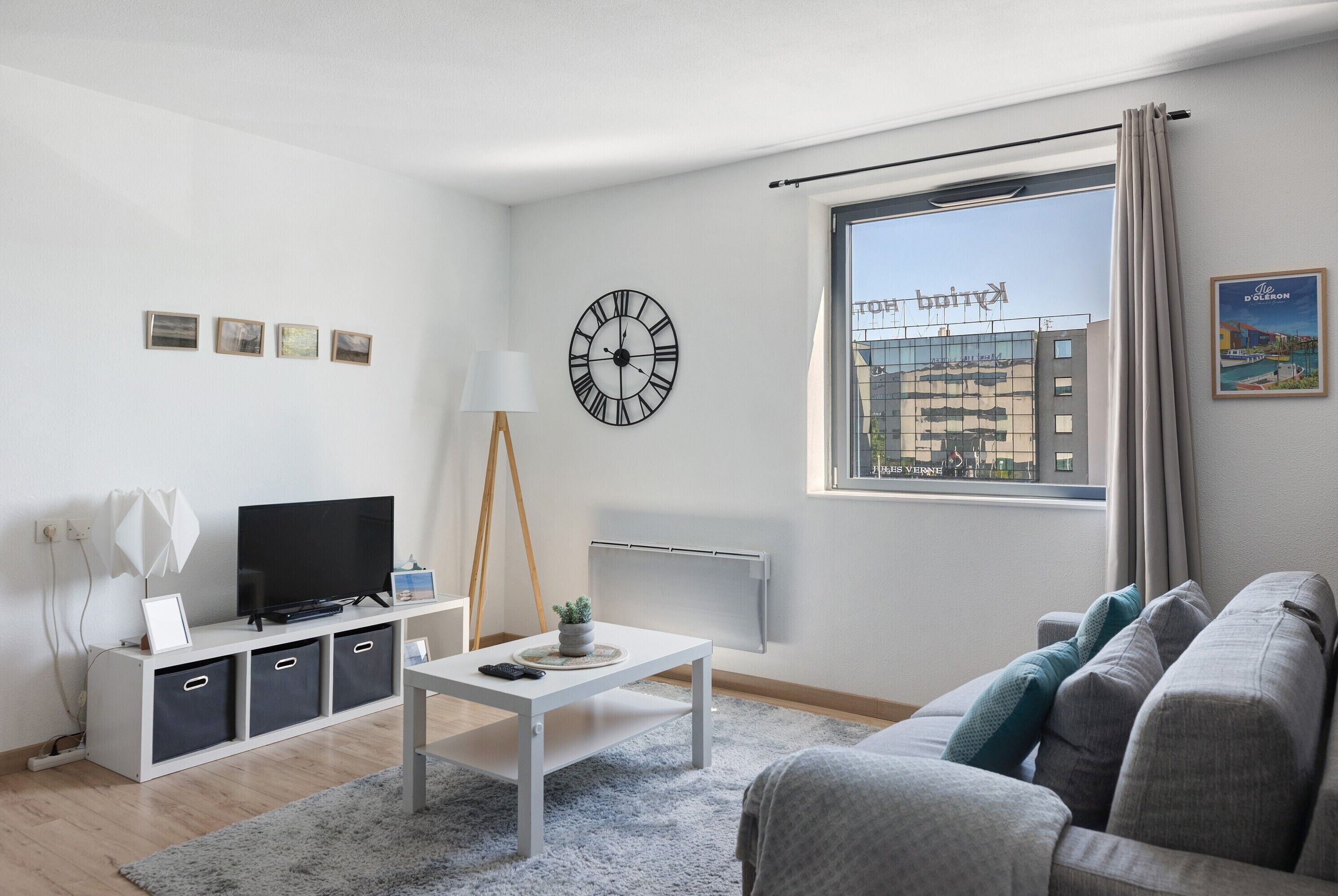 En moderne stue med et fjernsyn, et hvidt sofabord, en grå sofa og et stort vindue med udsigt til bygninger.