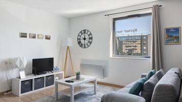 En moderne stue med et fjernsyn, et hvidt sofabord, en grå sofa og et stort vindue med udsigt til bygninger.