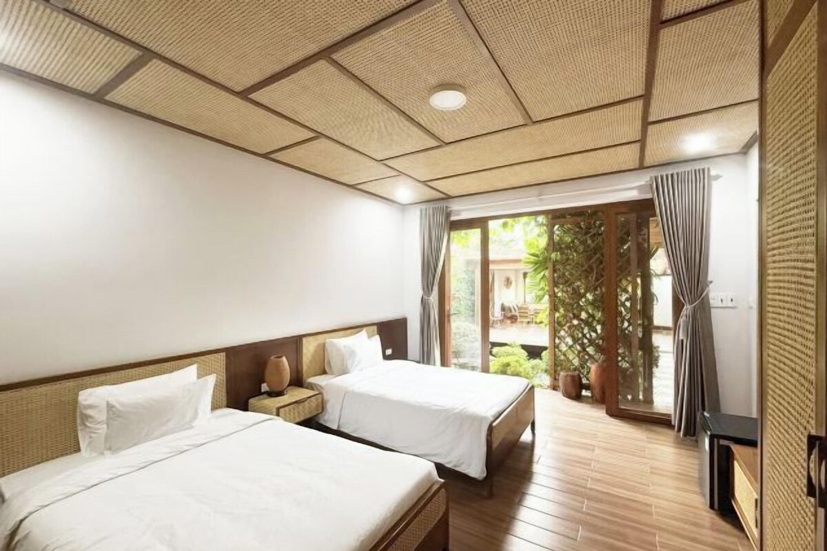 Een hotelkamer met twee bedden, een houten vloer, een groot raam met gordijnen en uitzicht op groen.