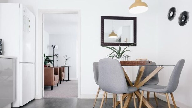 Et moderne køkken med spiseplads, med et glasbord med træben, grå polstrede stole og en hængende lampe.
