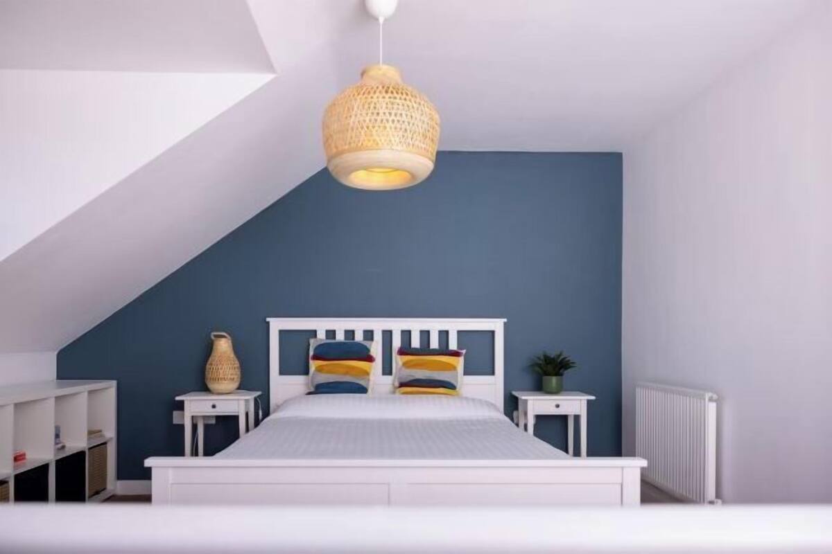 A bedroom with a sloped ceiling, a bed with a white headboard, a bedside table, a nightstand, and a hanging light fixture.