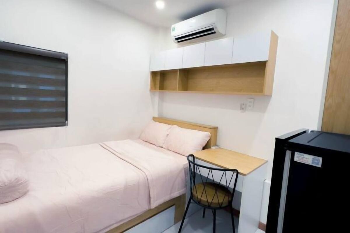 Une petite pièce comprenant un lit, un bureau, une chaise, un appareil de climatisation et un réfrigérateur noir.