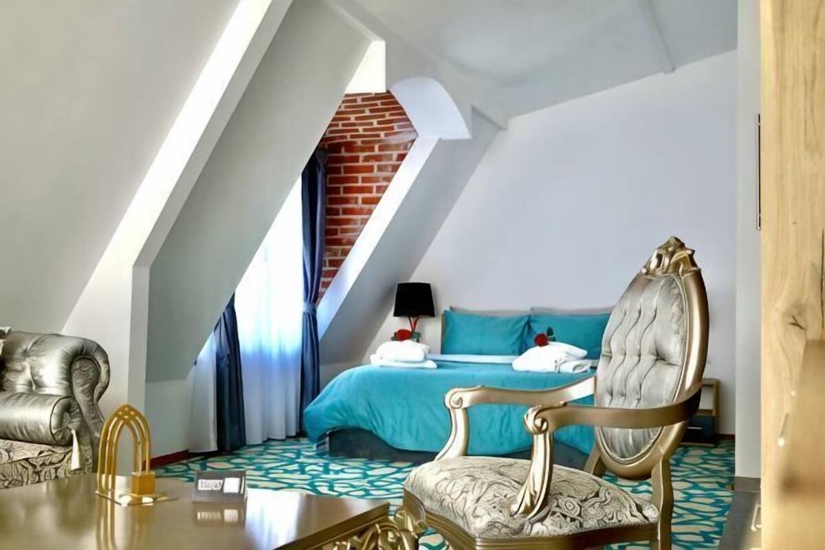 A room with a sloped ceiling, a bed with a blue bedspread, a sitting area with a patterned sofa, and a small table.