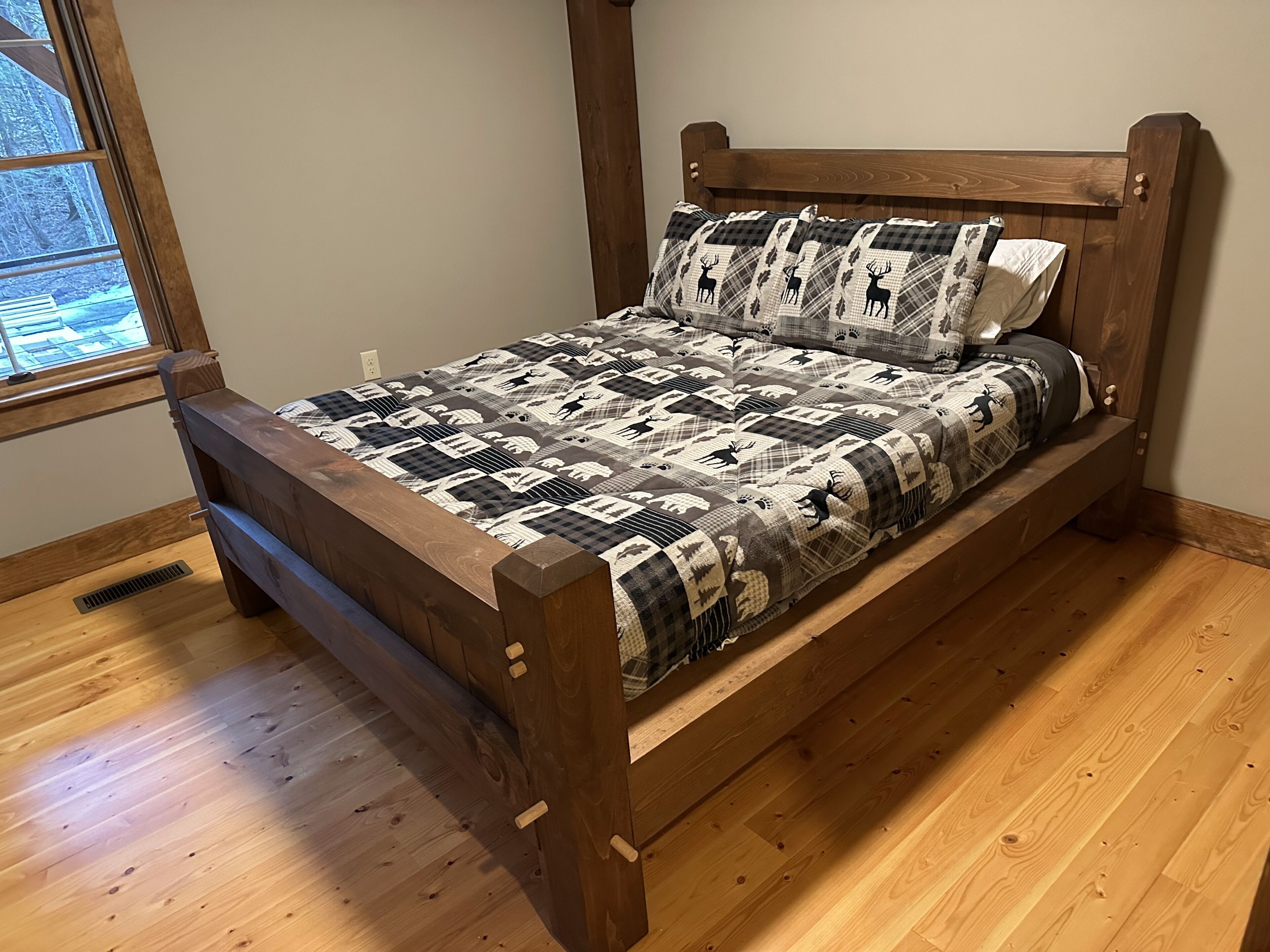 A wooden bed with a checkered quilt and matching pillows, in a room with wooden flooring and a window.