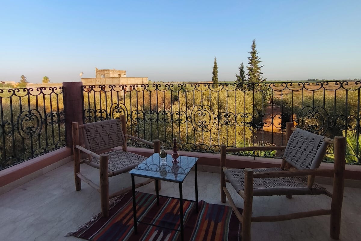 Ein Balkon mit zwei Holzstühlen, einem kleinen Tisch, einem gemusterten Teppich und einer Aussicht auf Bäume und Gebäude in der Ferne.