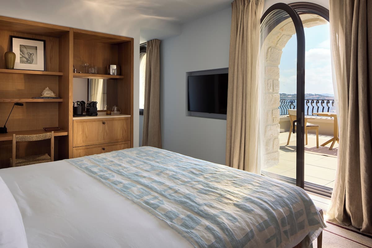 A hotel room with a bed, wooden shelving with books and a vase, a flat-screen TV, and a balcony with a view.