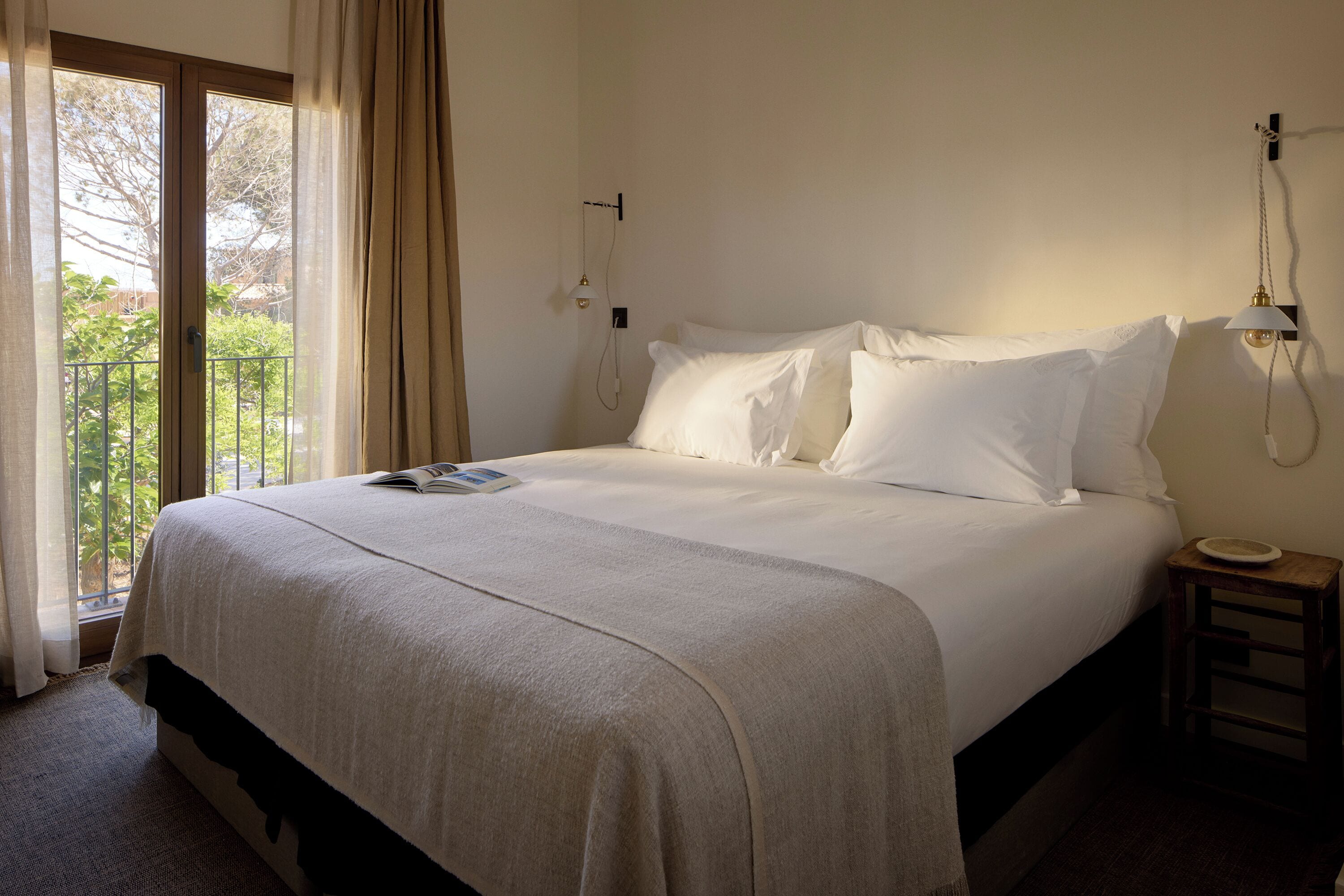 A neatly made bed with a white comforter and pillows, a bedside table with a lamp, and a view of greenery through a glass door.